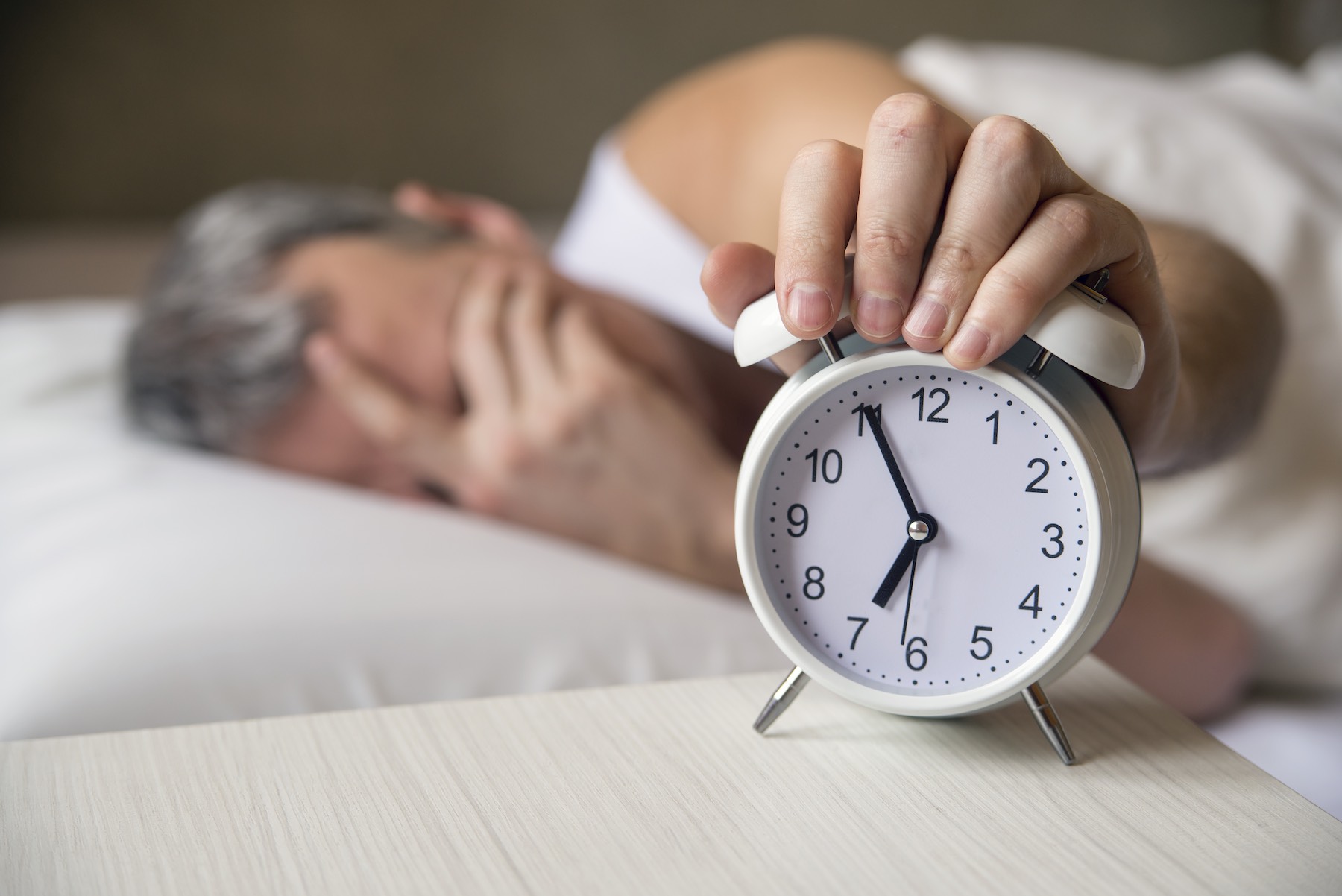Man lying in bed turning off an alarm clock in the morning at 7am. Attractive man sleeping in his bedroom. Annoyed man being awakened by an alarm clock in his bedroom